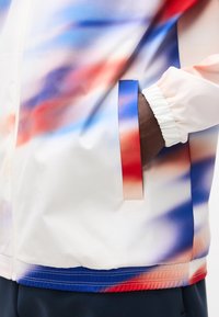 Χέρι στην τσέπη ενός πολύχρωμου μπουφάν με μπλε, κόκκινες και λευκές διαβαθμισμένες αποχρώσεις και ελαστικό μανίκι.