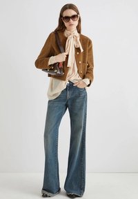 Giacca in velluto a coste marrone con bottoni dorati, blusa crema con nodo, jeans a zampa d'elefante blu, tiene una piccola borsa marrone e indossa occhiali da sole scuri.