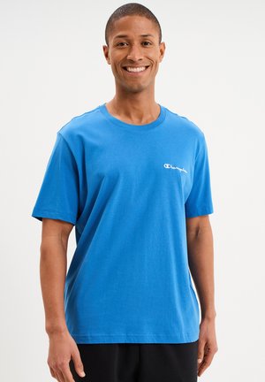 Lachende man met een blauw Champion T-shirt en zwarte broek, staand tegen een effen witte achtergrond.