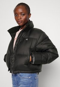 Svart puffjakke med høy krage, glidelåslommer og Calvin Klein-logo på brystet og ermene. Glatt, blank tekstur.