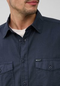 Marineblauw button-up shirt met een kraagontwerp, voorzien van twee voorzakken en een klein merklabel op de linkse zak. Glad stoffen textuur.