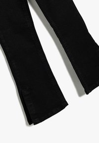 Svarta jeans med utsvängd ben, mjuk textur, utskärningar vid fållen och en klassisk fem-fickdesign. Slät yta och skräddarsydd passform.