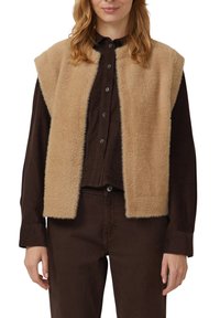 Beige mouwloze vest met een pluizige textuur, gedragen over een donkerbruine blouse met knopen. Eenvoudig ontwerp zonder zichtbare sluitingen of patronen.