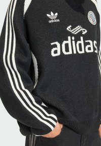 Svart stickad Adidas-tröja med en vit logotyp och tre vita ränder på varje ärm. Ribbstickade muddar och fåll, med tydlig textur.