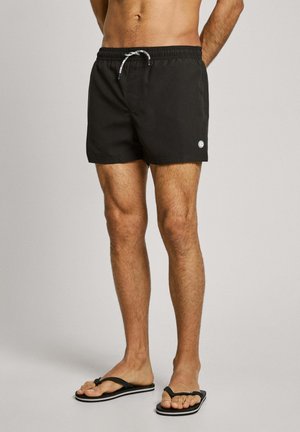 Schwarze Schwimmshorts mit elastischem Bund und Kordelzug. Besitzt ein kleines Logo am rechten Bein. Getragen mit schwarzen Flip-Flops.