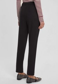 Pantaloni neri con vita elastica e gamba dritta, caratterizzati da una texture liscia e senza tasche visibili, abbinati a scarpe appiattite con borchie.