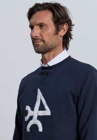 Sudadera azul marino con un logo blanco bordado en el centro, cuello redondo y tejido texturizado. Presenta un ajuste casual y mangas largas.