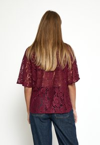 Top in pizzo bordeaux con maniche corte, design floreale stampato, texture semi-trasparente, indossato sopra pantaloni in denim scuro, visto da dietro.