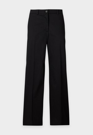 Femme aux cheveux courts et foncés, portant un haut bleu clair à manches courtes, un pantalon noir à jambes larges, et des talons pointus beiges, debout devant un fond uni.