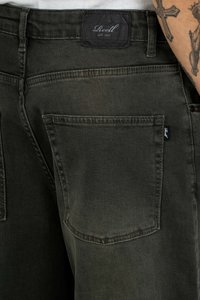Dark green denim jeans with a woven texture, featuring a stylish back pocket and a black label with white text above the waistband.