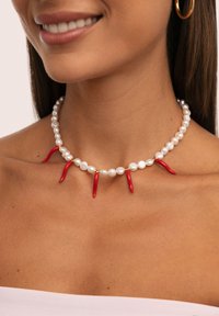 Primo piano di una donna sorridente che indossa una collana di perle con cinque pendenti a forma di peperoncino rosso e un orecchino a cerchio dorato.