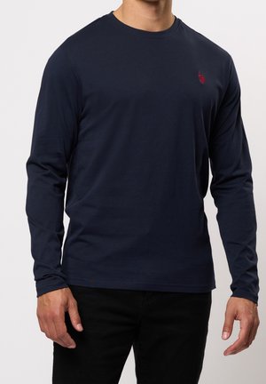 Homme portant une chemise bleu marine unie à manches longues avec un petit logo rouge brodé sur la poitrine gauche, associée à un pantalon noir.