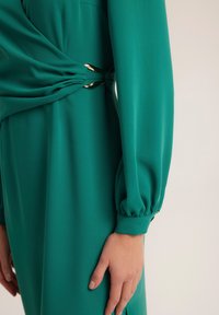 Turkusowa sukienka z długimi rękawami i marszczonymi mankietami, z twistowym zdobieniem w talii i metalowymi akcentami do zapięcia.