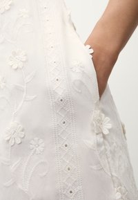 Robe blanche brodée présentant des motifs floraux, un tissu texturé, des coutures détaillées et une poche latérale avec des accents décoratifs.