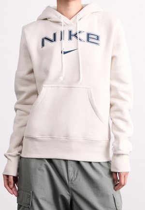 Personne portant un sweat à capuche Nike beige avec poche avant et cordons, associé à un pantalon cargo gris.