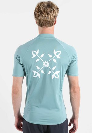 Homme tourné de dos portant une chemise bleu clair à manches courtes avec un motif floral blanc en forme de croix dans le dos.