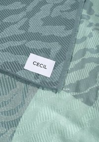 Tela texturizada en tonos verde azulado y verde menta que muestra un patrón geométrico, con una etiqueta tejida que dice "CECIL" en uno de los bordes.