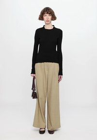 Black fitted long-sleeve top with ribbed texture, paired with beige wide-leg trousers. Model holds a small brown handbag and wears brown flats.
