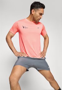 Herren-Sportoutfit mit einem leuchtend korallenfarbenen Mesh-T-Shirt mit kurzen Ärmeln und grauen, körpernahen Shorts. Enthält Marken- und Logodetails.