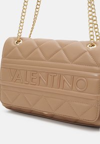 Quilted beige handbag with embossed "VALENTINO" logo. Features gold chain strap and smooth leather texture. Rectangular shape with subtle stitching.