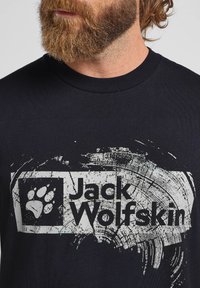 T-shirt in cotone nero con grafica circolare grigia che presenta il testo "Jack Wolfskin" e un simbolo della zampa, design con collo rotondo.