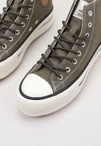 Zapatillas altas en lona verde oliva con puntera y suela de goma blanca, detalle de franja negra y parche de logo marrón en el lateral.