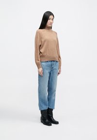 Καφές πλεχτός πουλόβερ με ζιβάγκο και ραβδωτές λεπτομέρειες, συνδυασμένο με ανοιχτό μπλε ψηλόμεσο τζιν και μαύρες μπότες μέχρι τον αστράγαλο.