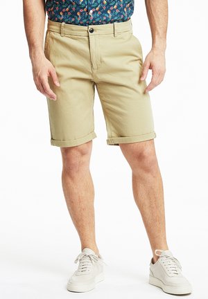 Mand iført beige opkørte shorts, hvide sneakers og en blå skjorte med blomsterprint, stående mod en hvid baggrund.