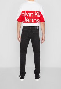 Calvin Klein white and red graphic t-shirt with block lettering, paired with fitted black jeans and black footwear, viewed from the back.