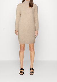 Vestito beige a collo alto realizzato in morbido tessuto a maglia, con maniche lunghe e orlo a costine. Abbinato a sandali neri con cinturini.
