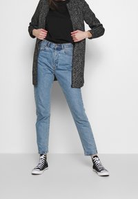 Chaqueta negra texturizada, camiseta negra, jeans de cintura alta de color azul claro y zapatillas deportivas negras de caña alta, con cordones de contraste y un corte holgado.
