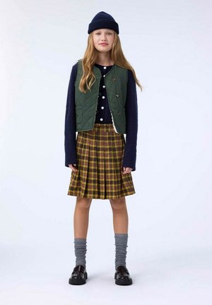 Gilet trapuntato verde sopra un maglione a maniche lunghe blu scuro, sovrapposto a una gonna plissettata giallo a quadri. Calze grigie fino al ginocchio e scarpe nere.