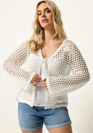 Femme blonde portant un cardigan en crochet maille ouverte blanc par-dessus un top blanc et un short en denim bleu, debout avec les mains près des liens avant.