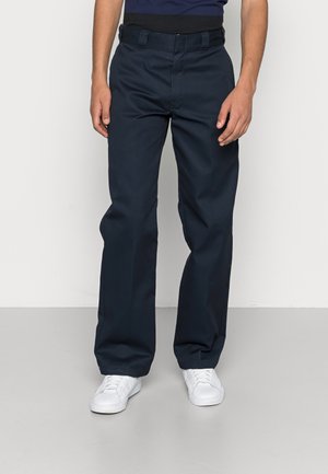 Man draagt marineblauwe rechte broek en witte sneakers, staand tegen een effen lichtgrijze achtergrond.