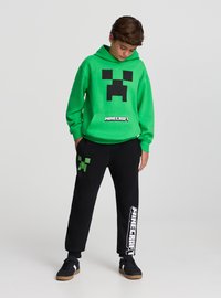 Felpa verde con cappuccio e grafica nera del volto di Creeper, abbinata a pantaloni della tuta neri con icona verde di Creeper e scritta bianca "Minecraft".