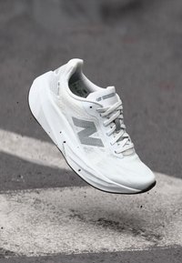 Chaussure de sport blanche avec une tige en maille, surface texturée et accents gris. Présente un orteil arrondi et une semelle légère et rembourrée.