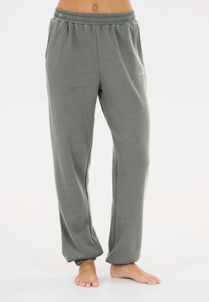 Person iført grå sweatpants med elastik i taljen står barfodet på en ensfarvet hvid baggrund.