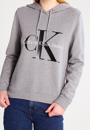 Sweatshirt gris avec capuche, présentant un grand logo noir "CK" et le texte "Calvin Klein Jeans". Fabriqué en matériau doux, avec des poignets côtelés.
