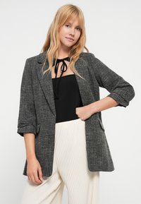 Blazer cinzento texturizado com corte solto, lapelas com entalhes e mangas arregaçadas. Usado por cima de uma blusa preta e combinado com calças plissadas de cor clara.