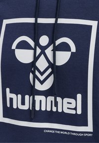 Marineblauer Stoff mit einem weißen stilisierten Bienenlogo und dem Text "hummel" innerhalb eines Quadrats, mit Kordeln vorne und einem Slogan darunter.