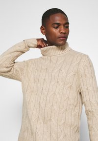 Suéter de punto beige con cuello alto que presenta un patrón de diamante texturizado y puños acanalados, destacando un ajuste cómodo y relajado.