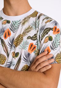 Kortärmad t-shirt med en vit bas som har ett tropiskt frukt- och palmbladsmönster, inklusive ananaser och papayas, med en mjuk textur.