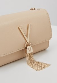 Pochette beige strutturata con superficie testurizzata, prominente logo "V" dorato e dettaglio a nappina con catena, dotata di chiusura e rifiniture con cuciture.