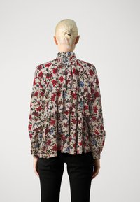 Blomstret bluse med en flæset design, der har røde, blå og grønne mønstre på en beige stof. Høj krave og lange ærmer.