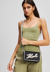 Black textured crossbody bag with white lettering "Hol." Features a removable strap and a rectangular shape, designed for casual use.