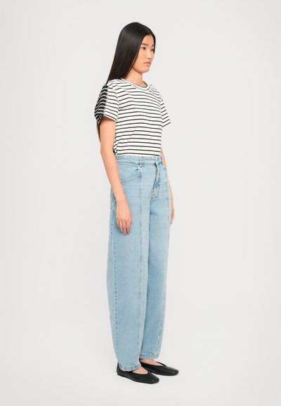 Woman with long straight hair wearing a black-and-white striped shirt, light blue high-waisted jeans, and black flats, standing sideways.