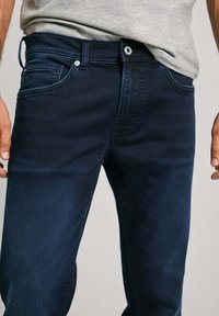 Tumed sinised teksad, millel on keha järgi iste, klassikaline viie taskuga stiil ja peened õmblusdetailid õmbluste ja taskute ääres.