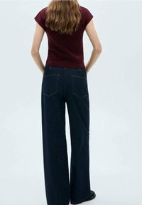 Jeans de pierna ancha en azul oscuro con bolsillos traseros, combinados con una blusa ajustada de color burdeos. El material parece suave y tiene una textura lisa.
