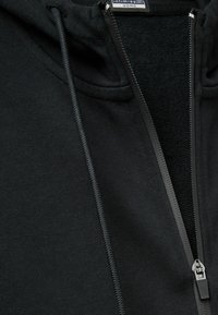 Sudadera con cremallera negra, con un forro interior texturizado, cordones planos y una cremallera suave con un tirador metálico.