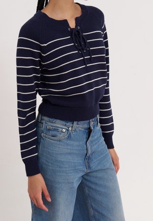 Femme portant un pull bleu marine avec des rayures horizontales blanches et un détail noué au niveau du col, associé à un jean bleu taille haute.
